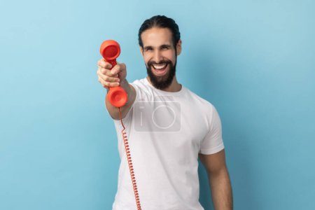 Téléchargez les photos : Portrait d'un bel homme heureux avec barbe portant un T-shirt blanc tenant et montrant un téléphone rétro combiné à la caméra, demandant à répondre au téléphone. Studio intérieur tourné isolé sur fond bleu. - en image libre de droit