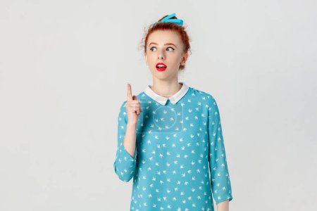 Photo for Portrait of excited clever smart red haired woman with bun hairstyle, standing raised index finger up, having idea, looking away, wearing blue dress. Indoor studio shot isolated on gray background. - Royalty Free Image