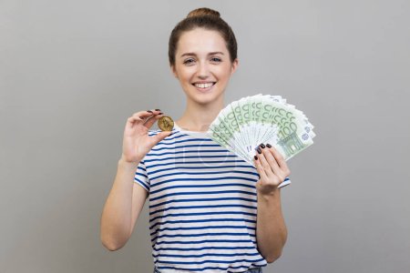 Photo for Portrait of delighted positive woman wearing striped T-shirt holding golden coin of crypto currency and big fan of euro banknotes. Indoor studio shot isolated on gray background. - Royalty Free Image
