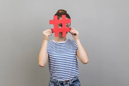 Téléchargez les photos : Portrait de femme inconnue portant un T-shirt rayé regardant à travers le signe rouge hashtag, à la recherche de messages appropriés dans les médias sociaux, espionnage. Studio intérieur tourné isolé sur fond gris. - en image libre de droit