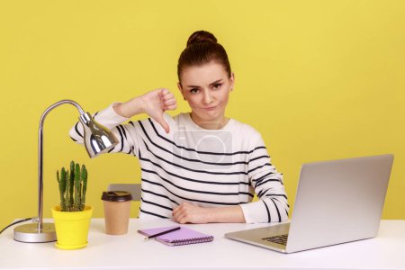 Photo for Displeased woman office worker showing thumbs down, dislike gesture, expressing disapproval, criticizing sitting at workplace with laptop. Indoor studio studio shot isolated on yellow background. - Royalty Free Image