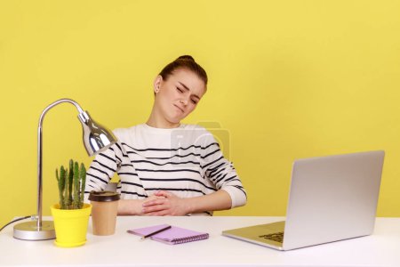 Photo for Unhappy woman office manager sitting at workplace, feeling discomfort and abdominal cramps, suffering heartburn or dysmenorrhea. Indoor studio studio shot isolated on yellow background. - Royalty Free Image