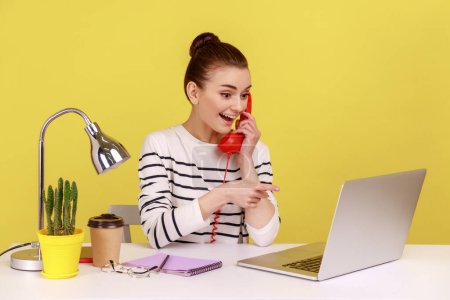 Photo for Woman secretary making calls on landline telephone working on laptop sitting at office, looking and pointing at notebook display. Indoor studio studio shot isolated on yellow background. - Royalty Free Image