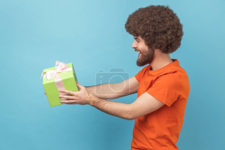 Photo for Side view of man with Afro hairstyle wearing orange T-shirt giving gift, congratulating on birthday and offering surprise, holiday bonus wrapped in box. Indoor studio shot isolated on blue background. - Royalty Free Image