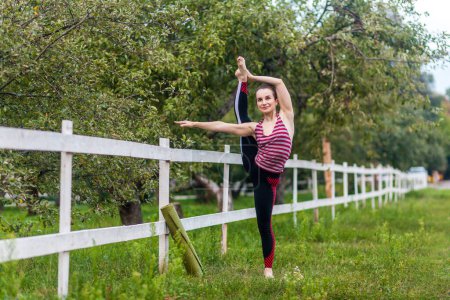 Photo for Full length portrait of flexible strong young girl stretching her leg, practicing yoga, having workout outdoor, raising leg in air, doing split stretch one foot, looking smiling away. - Royalty Free Image