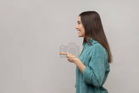 Photo for Side view of happy beautiful young adult woman standing, pointing and showing copyspace with happy face and smile, wearing casual style jacket. Indoor studio shot isolated on gray background. - Royalty Free Image