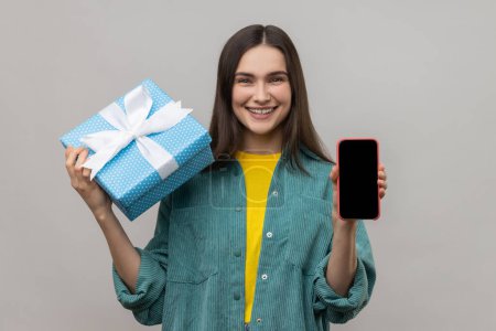 Photo for Present, bonus for mobile user. Woman holding gift box and cell phone with empty display for online shopping advertising, wearing casual style jacket. Indoor studio shot isolated on gray background. - Royalty Free Image