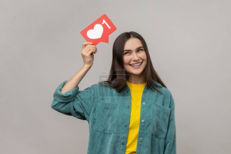 Photo for Portrait of smiling young woman holding heart icon over head, looking at camera, share blog with trendy content, wearing casual style jacket. Indoor studio shot isolated on gray background. - Royalty Free Image