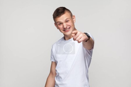 Foto de Retrato de un adolescente riéndose feliz vistiendo una camiseta mirando a la cámara con una sonrisa dentada, apuntando a la cámara con el dedo, riéndose de ti. Estudio interior plano aislado sobre fondo gris. - Imagen libre de derechos