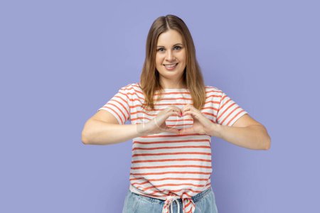 Photo for Portrait of blond woman wearing striped T-shirt holding hands in shape of heart showing romantic gesture, love confession, valentines day celebration. Indoor studio shot isolated on purple background. - Royalty Free Image