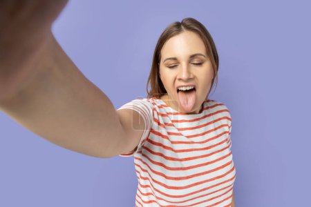 Photo for Portrait of funny childish woman in striped T-shirt taking selfie picture, point of view, showing tongue out to camera, making front selfportrait. Indoor studio shot isolated on purple background. - Royalty Free Image
