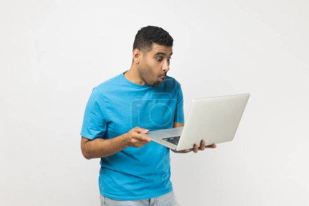 Téléchargez les photos : Portrait d'un homme étonné et non rasé portant un T-shirt bleu tenant un ordinateur portable dans les mains, travaillant en ligne, regardant l'écran avec de grands yeux. Studio intérieur tourné isolé sur fond gris. - en image libre de droit
