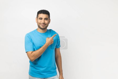 Foto de Retrato de sonriente alegre hombre sin afeitar atractivo usando azul camiseta de pie apuntando espacio de copia para publicidad o texto promocional. Estudio interior plano aislado sobre fondo gris. - Imagen libre de derechos