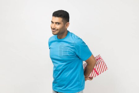 Photo for Portrait of positive cheerful unshaven man wearing blue T- shirt standing with present box behind back, gift for holiday, congratulating. Indoor studio shot isolated on gray background. - Royalty Free Image