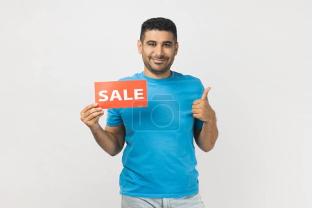 Photo for Portrait of cheerful joyful man wearing blue T- shirt standing holding sale card in hands looking at camera with toothy smile, showing thumb up. Indoor studio shot isolated on gray background. - Royalty Free Image