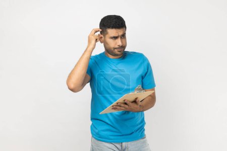 Photo for Portrait of pensive puzzled unshaven man wearing blue T- shirt standing with clipboard, writing something on paper, thinking about plan. Indoor studio shot isolated on gray background. - Royalty Free Image