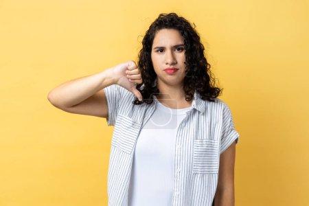 Photo for Portrait of woman with dark wavy hair criticizing bad quality with thumbs down displeased grimace, showing dislike gesture, expressing disapproval. Indoor studio shot isolated on yellow background. - Royalty Free Image