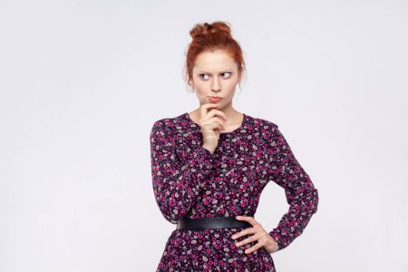 Photo for Portrait of redhead woman wearing dress holding chin and pondering idea, planning strategy, musing and solving difficult question in mind. Indoor studio shot isolated on gray background. - Royalty Free Image