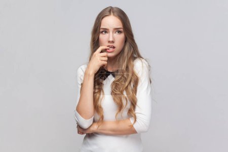 Portrait of nervous beautiful young adult woman with long blond hair biting her finger, looking away, thinking about something important. Indoor studio shot isolated on gray background.