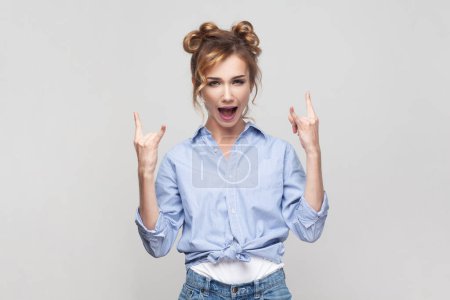 Photo for Portrait of blonde woman showing rock and roll gesture heavy metal sign enjoying favorite music on party has fun, exclaims from joy, wearing blue shirt. Indoor studio shot isolated on gray background. - Royalty Free Image