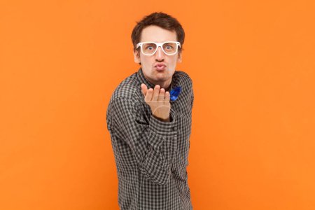 Foto de Retrato del gracioso hombre positivo nerd enviando besos al aire, enamorándose, coqueteando, vistiendo camisa con pajarita azul y gafas blancas. Estudio interior plano aislado sobre fondo naranja. - Imagen libre de derechos