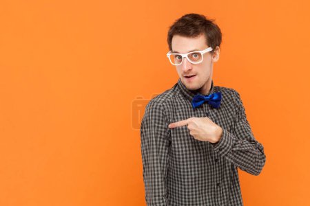 Foto de Retrato del hombre nerd apuntando lejos en el espacio de copia, discute promo increíble, da paso o dirección, usando camisa con pajarita azul y gafas blancas. Estudio interior plano aislado sobre fondo naranja. - Imagen libre de derechos