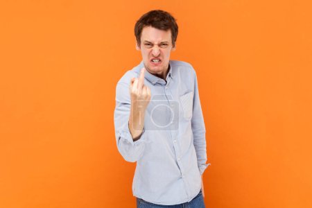 Foto de Portrait of crazy man shows fuck you sign looking with poker face being vulgar and has quarrel with someone, wearing light blue shirt. Indoor studio shot isolated on orange background. - Imagen libre de derechos