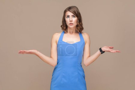 Photo for Portrait of puzzled confused young adult woman with wavy hair standing shrugging shoulders, don't know what to do, wearing blue dress. Indoor studio shot isolated on light brown background. - Royalty Free Image