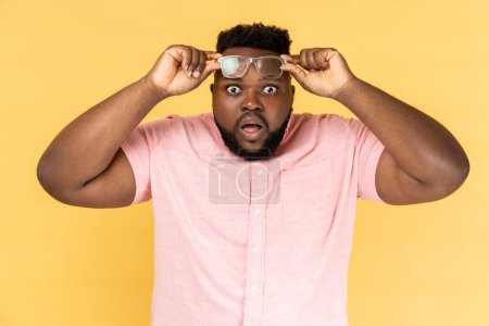 Photo for Portrait of shocked amazed surprised man wearing pink shirt standing raised his optical glasses and looking at camera with big eyes and open mouth. Indoor studio shot isolated on yellow background. - Royalty Free Image