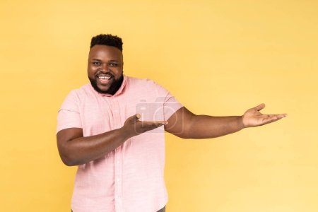 Photo for Please, take for free. Portrait of man wearing pink shirt welcoming with wide open arms and smiling kindly, looking at camera with toothy smile. Indoor studio shot isolated on yellow background. - Royalty Free Image