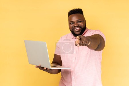 Téléchargez les photos : Portrait d'un homme barbu optimiste portant une chemise rose tenant un ordinateur portable, pointant du doigt la caméra et souriant, vous choisissant, annonce d'emploi indépendant. Studio intérieur tourné isolé sur fond jaune - en image libre de droit