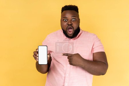 Photo for Portrait of shocked man wearing pink shirt pointing at cell phone screen, looking at camera with big eyes, showing mobile phone with white display. Indoor studio shot isolated on yellow background. - Royalty Free Image
