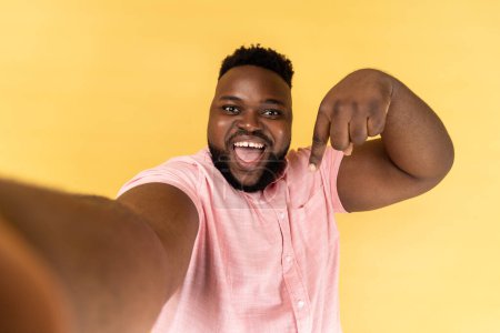 Photo for Portrait of happy joyful man blogger wearing pink shirt making point of view photo, having friendly excited , POV pointing down, asking to subscribe. Indoor studio shot isolated on yellow background - Royalty Free Image