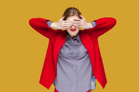 Photo for Portrait of shocked woman with red lips standing covering her eyes with palm, do not want to see something scary, wearing red jacket. Indoor studio shot isolated on yellow background. - Royalty Free Image