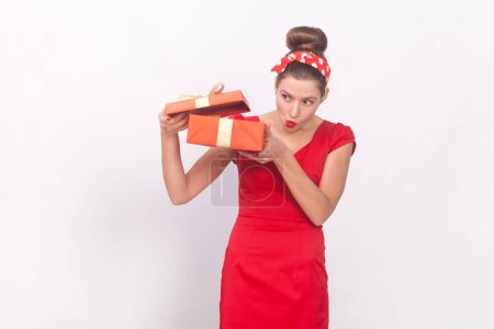 Photo for Portrait of curious attractive woman wearing red dress and head band holding present box, looking inside with interest, having gift on her birthday. Indoor studio shot isolated on gray background. - Royalty Free Image