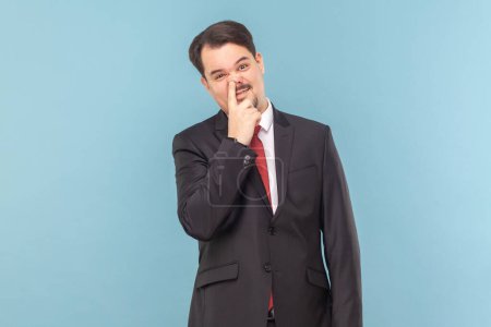Photo for Portrait of funny childish man with mustache standing having comical facial expression, keeps finger in nose, wearing black suit with red tie. Indoor studio shot isolated on light blue background. - Royalty Free Image