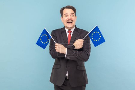 Téléchargez les photos : Homme souriant largement et tenant des drapeaux de l'Union européenne, symbole de l'Europe, association et communauté de l'UE, portant un costume noir avec cravate rouge. Studio intérieur tourné isolé sur fond bleu clair. - en image libre de droit