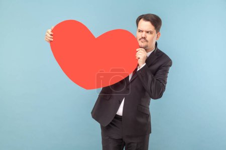 Photo for Portrait of confused cunning man with mustache standing with big red heart in hands, looking away, wearing black suit with red tie. Indoor studio shot isolated on light blue background. - Royalty Free Image