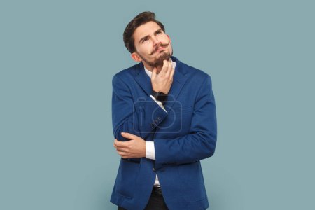 Photo for Portrait of thoughtful serious businessman with mustache standing and thinking about his work, holding his chin, wearing official style suit. Indoor studio shot isolated on light blue background. - Royalty Free Image