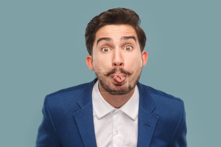 Photo for Portrait of funny handsome man with mustache standing sticking tongue out, showing childish behavior, wearing white shirt and jacket. Indoor studio shot isolated on light blue background. - Royalty Free Image