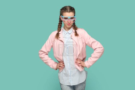 Foto de Retrato de chica adolescente divertida y confiada con trenzas que usan chaqueta rosa de pie con las manos en las caderas, usando dos gafas ópticas, eligiendo el mejor color. Estudio interior plano aislado sobre fondo verde - Imagen libre de derechos