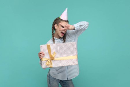 Photo for Portrait of joyful beautiful teenager girl wearing striped shirt and party cone, holding present box, showing v sign, celebrating her birthday. Indoor studio shot isolated on green background. - Royalty Free Image