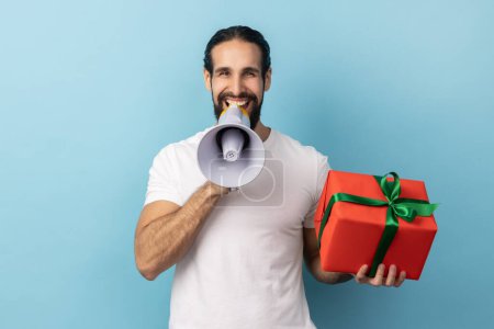 Téléchargez les photos : Portrait d'un bel homme positif avec une barbe portant un T-shirt blanc tenant une boîte cadeau rouge et criant en mégaphone, faisant une annonce. Studio intérieur tourné isolé sur fond bleu. - en image libre de droit