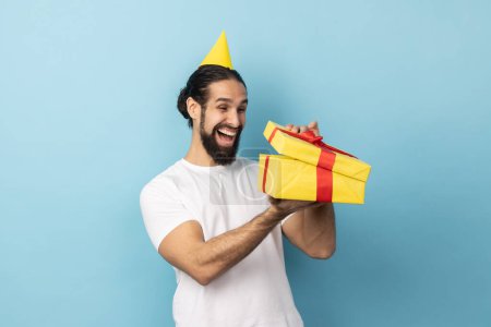 Photo for Portrait of extremely happy man wearing white T-shirt and party cone looking into gift box, opening present and peeking inside with happiness. Indoor studio shot isolated on blue background. - Royalty Free Image