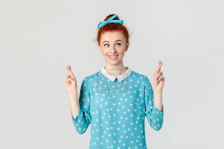 Photo for Portrait of smiling attractive hopeful ginger woman with bun hairstyle, standing with crossed fingers, making wish, wearing blue dress. Indoor studio shot isolated on gray background. - Royalty Free Image