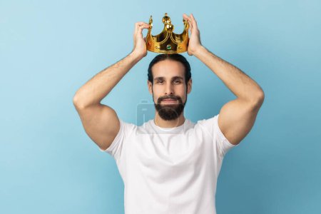 Photo for Portrait of serious independent man with beard in white T-shirt wearing golden crown, looking with arrogance and confidence, privileged status. Indoor studio shot isolated on blue background. - Royalty Free Image