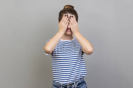 Photo for Don't want to look at this. Portrait of confused woman wearing striped T-shirt covering eyes with hand, feeling shamed and scared to watch. Indoor studio shot isolated on gray background. - Royalty Free Image