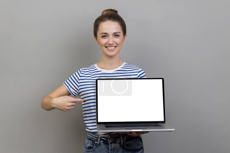 Téléchargez les photos : Portrait de femme portant un T-shirt rayé pointant du doigt sur un ordinateur portable avec un écran blanc vierge, montrant un écran vide, se vantant avec une publicité sur Internet. Studio intérieur tourné isolé sur fond gris - en image libre de droit