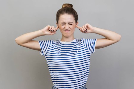 Photo for I dont want to hear this. Annoyed frustrated woman wearing striped T-shirt closing ears with fingers ignoring information, tired of irritating noise. Indoor studio shot isolated on gray background. - Royalty Free Image
