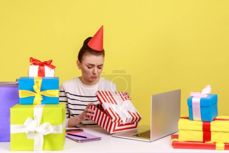 Photo for Unhappy woman in party cone sitting in office workplace, opening gift box and looking inside with sad upset expression, unwrapping present. Indoor studio studio shot isolated on yellow background - Royalty Free Image